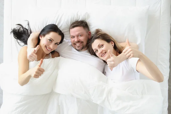 Två leende kvinnor och man ligger på sängen och håller tummarna upp — Stockfoto