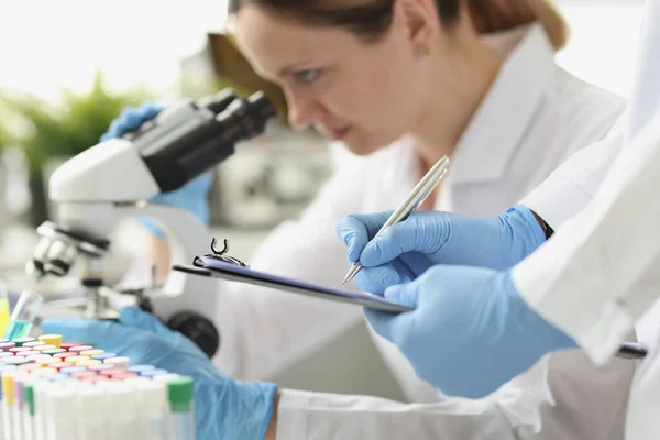 Γυναίκα χημικός κοιτάζοντας μέσα από μικροσκόπιο στο εργαστήριο. Βοηθός γραφής δεδομένων σε χαρτί — Φωτογραφία Αρχείου