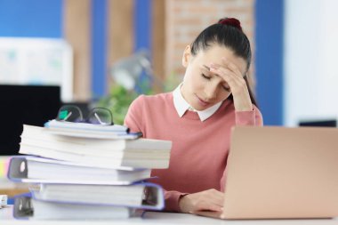 Dizüstü bilgisayarın arkasında oturan yorgun bir kadın ve bir sürü belge ve başını tutuyor.