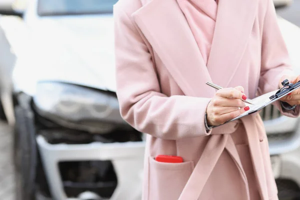Жінка в пальто заповнює документи на тлі розбитого автомобіля крупним планом — стокове фото