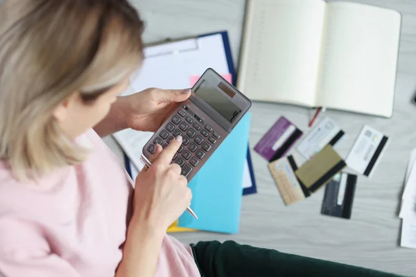 Mujer sentada en el piso cerca de tarjetas de banco dispersas y contando con la calculadora — Foto de Stock