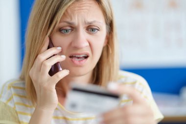 Cep telefonuyla konuşan ve elinde banka kartı tutan şaşırmış bir kadın.