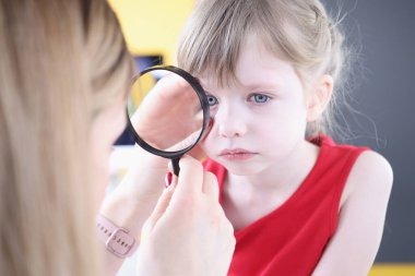 Doctor examines eye of little girl through magnifying glass closeup clipart