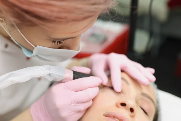 Master in beschermende medische masker maken van permanente oog make-up aan cliënt in schoonheidssalon — Stockfoto