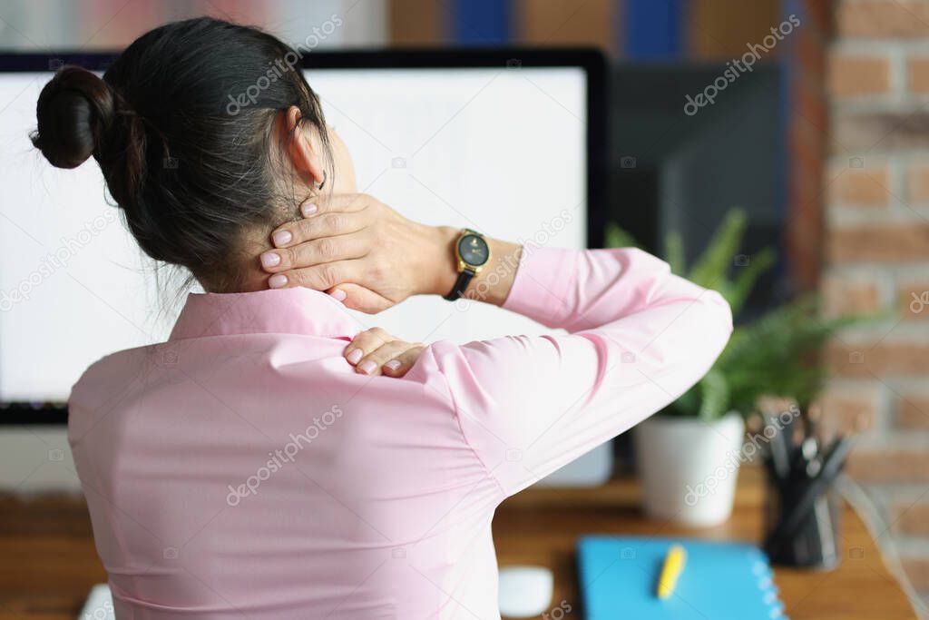 Young woman sitting at computer and holding her sore neck with her hand back view