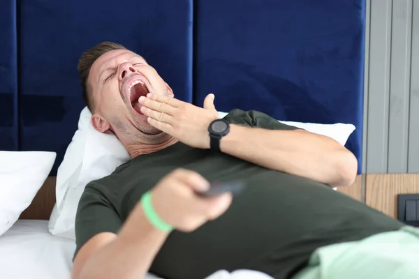 Jeune homme couché au lit avec télécommande à la main et bâillement — Photo