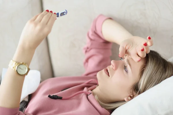 Больная молодая женщина лежит на диване и смотрит на термометр в руках — стоковое фото