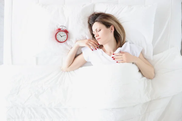 Ung kvinna ligger i sängen nära röd väckarklocka ovanifrån — Stockfoto