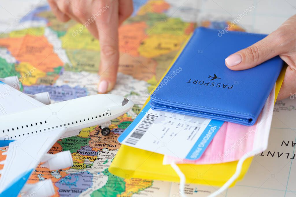 In hand a passport with airplane ticket and medical protective mask, finger points to country on world map
