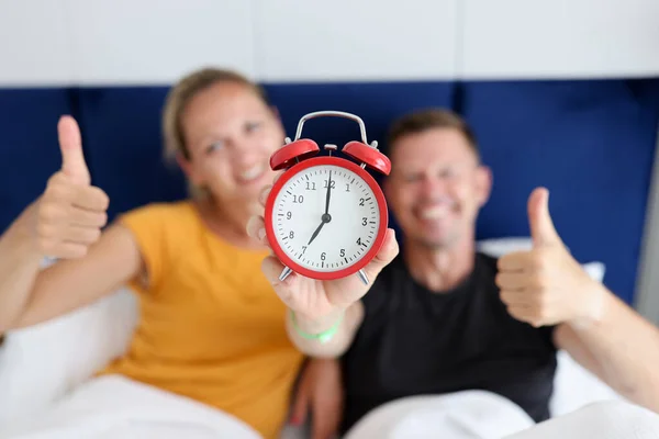 Jong en gelukkig man en vrouw met rode wekker en duimen omhoog terwijl liggen in de slaapkamer — Stockfoto