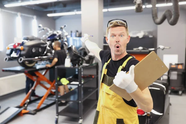 Здивований чоловік-ремонтник тримає кишеню з документами і вказує палець на мотоцикл — стокове фото