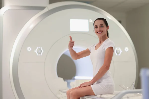 Young woman sitting on magnetic resonance imaging machine and showing thumb up — Stock Photo, Image