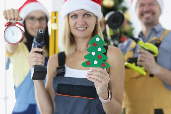 Grupo de reparadores de capataces en sombreros de Santa Claus sosteniendo taladro y despertador cerca del primer árbol de navidad —  Fotos de Stock