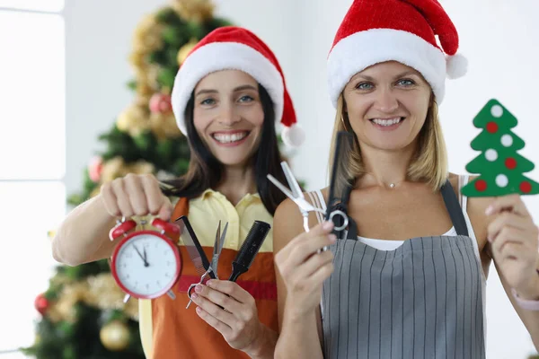 Dos mujeres sonrientes peluqueras en uniformes y sombreros de Santa Claus sostienen las tijeras del reloj despertador con peine. —  Fotos de Stock