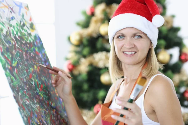 Sorridente artista donna in cappello di Babbo Natale disegna immagine sullo sfondo dell'albero di Natale — Foto Stock