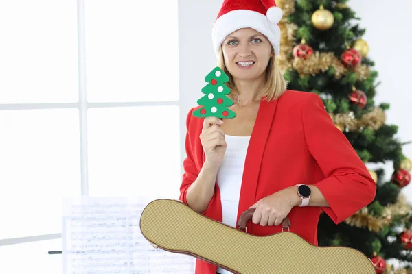 Noel Baba şapkalı gülümseyen kadın Noel ağacının arka planında gitar tutuyor. — Stok fotoğraf