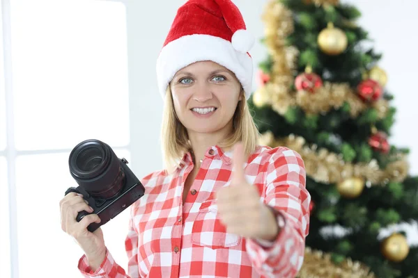 Vrouw fotograaf in santa claus hoed op achtergrond van kerst boom maakt duimen omhoog gebaar — Stockfoto