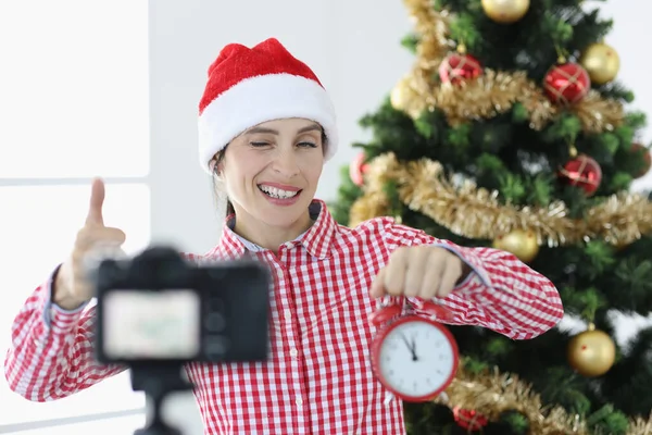 Noel Baba şapkalı genç kadın blogcu çalar saati tutuyor ve kameraya göz kırpıyor — Stok fotoğraf