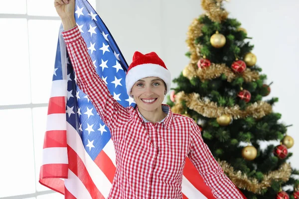 Vreugdevolle vrouw in kerstman hoed houdt vlag van Amerika tegen de achtergrond van Nieuwjaar boom — Stockfoto