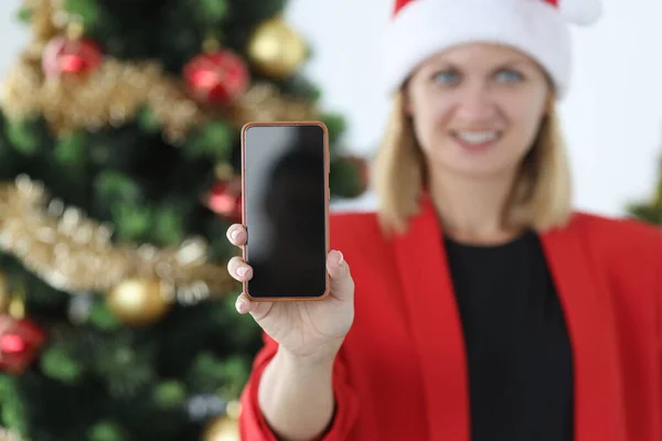 Mujer sonriente en sombrero de Santa Claus sostiene muestra teléfono inteligente en el fondo del árbol de Navidad primer plano — Foto de Stock