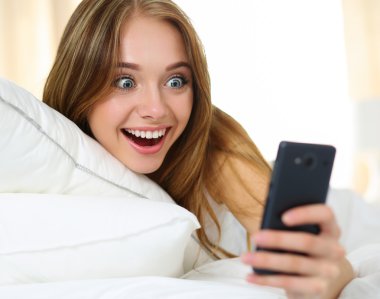 Young beautiful blonde woman lying in bed and holding cellphone clipart