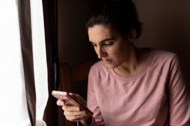 ciddi bir kadın cep telefonu kullanıyor doğal ışığı olan bir pencerenin yanında.