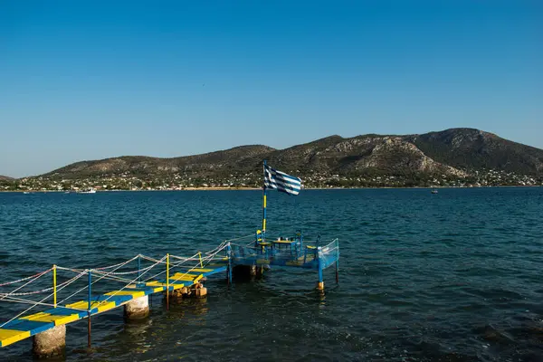 一个希腊国旗在海水上方的木制平台上飘扬 导致夏天在海滩旁边有一张桌子和三张椅子共进晚餐 — 图库照片