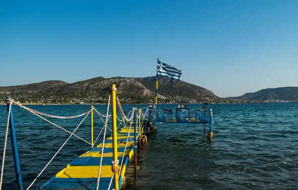 一个希腊国旗在海水上方的木制平台上飘扬 导致夏天在海滩旁边有一张桌子和三张椅子共进晚餐 — 图库照片