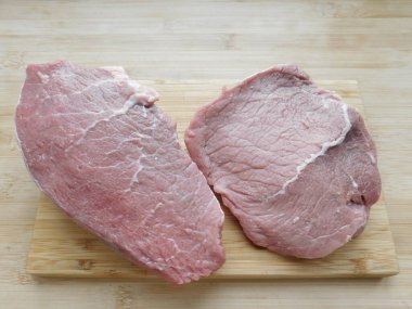 Ahşap bir masanın üzerindeki tahta kesme tahtasının üzerinde iki domuz bifteği. Et yemekleri için malzemeler.