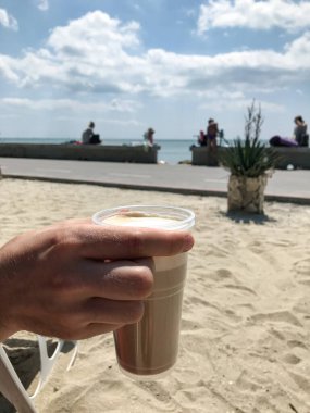 Bir kadının eli plajda plastik bir bardakta kapuçino tutuyor. Balchik. Bulgaristan
