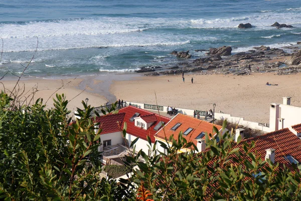 coastal village on the ocean from the bird\'s eye view directly on the beach and people with the view over green bushes down to the village and beach and the ocean to the horizon and blue, cloudless sky