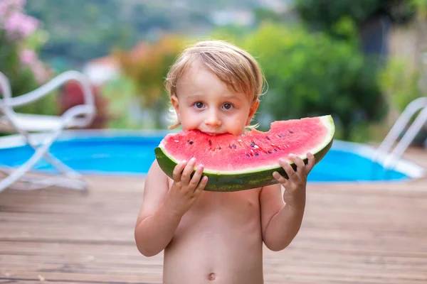 Toddler child eating watermelon near swimming pool during summer holidays. Kids eat fruit outdoors. Healthy snack for children. Little boy playing in the garden holding a slice of water melon.