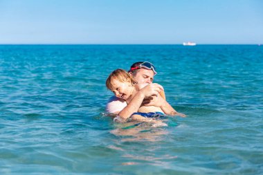 Baba ve küçük oğlu yaz tatillerinde deniz suyunda birlikte yüzerek ve oynayarak eğleniyorlar. Aile bağı, birliktelik. Deniz kenarı tatili.