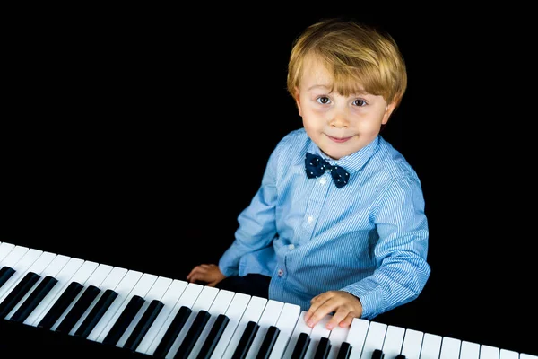 エレガントなシャツと蝶ネクタイを身に着けている愛らしい小さな幼児の少年の肖像画を閉じます 笑顔の子供は黒の背景に隔離されたピアノに座っている 子供の遊び音楽 — ストック写真