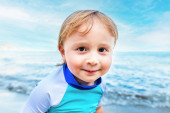 Közelkép egy kisfiúról, aki hároméves, napvédő pólóban van a tengerparton, közel a tengerhez. A gyermekek nyaralása a tengerparton.