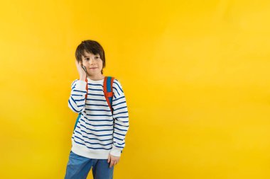 A young boy of nine years old with a backpack on his shoulders talking on his  smartphone with his parents or friends, isolkated on yellow background, copy space on the right.