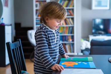 Küçük çocuk masada oturuyor, Coronavirus karantinası sırasında evde öğreniyor. Bebecik, erken gelişim kitabına bakıyor. Çıkartmalarla oynamada çocuk eğitimi.