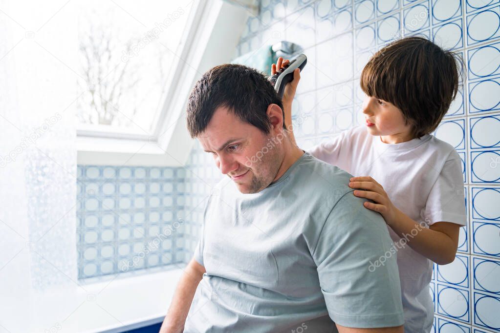 Young boy helping his father to cut hair at home. Son shaving daddy with clipper staying in bathroom during coronavirus quarantine. Hairdressing salons closed.
