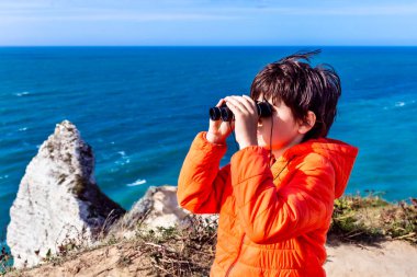 Turuncu ceketli bir çocuk, kayanın üzerinde duruyor ve dürbünle okyanusa bakıyor. Etretat Aval uçurumu, kayalar ve doğal kemer işaretleri. Normandiya, Fransa.