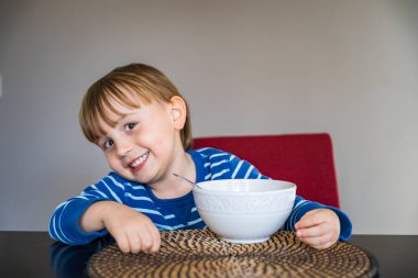 Evde yemek yiyen sevimli küçük bir çocuk. Bebek masada oturuyor, kasede çorba tadıyor. Mutlu gülümseyen çocuk sağlıklı ev yemekleri yiyor. Uzayı kopyala, taklit et