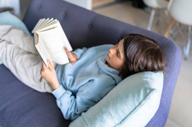 Açık mavi kazak giyen, evdeki kanepede uzanmış, faiziyle kitap okuyan genç bir çocuk..