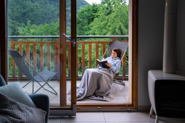 Fransa 'daki dağlarda terasta bir iskemlede oturan çocuk kitap okuyor. Çocuk battaniyeyle örtülmüş, açık havada. Pencereden bak