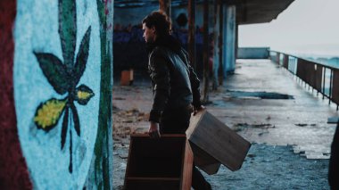 Deri ceketli adam eski mobilyaları kenevir etiketinin yanına sürüklüyor. 