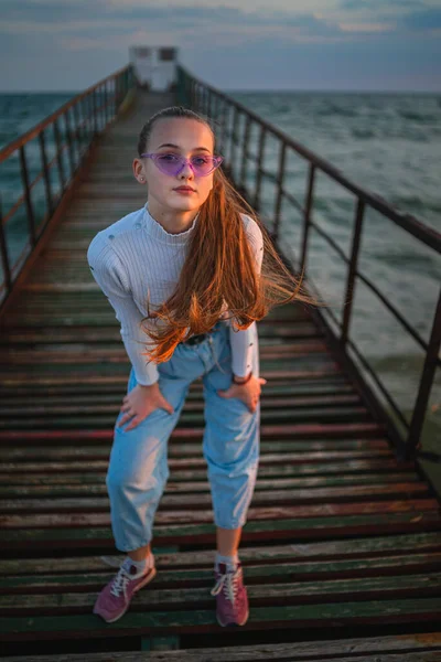 Aranyos lány rózsaszín szemüvegben pózol a mólón a tengeren naplementekor — Stock Fotó