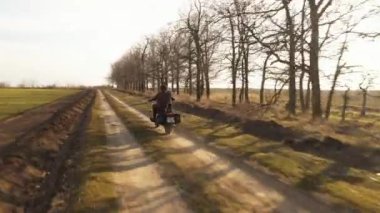 Deri takım elbiseli beyaz bir adam engebeli arazide motosiklet sürüyor.