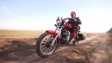 Yakışıklı adam tarladan geçen toprak yolda kırmızı bir kafe motosikleti sürüyor.