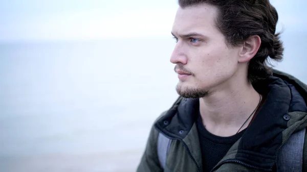 Attrayant jeune homme aux yeux bleus avec barbe et cheveux bouclés portrait solitaire sur la côte Photo De Stock