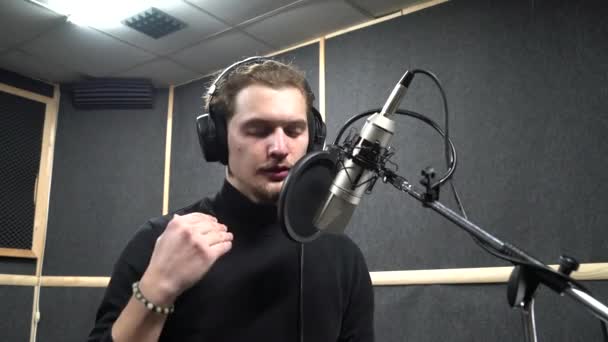 Krullend jong in hoofdtelefoon zingt in de buurt van microfoon in voice recording studio — Stockvideo