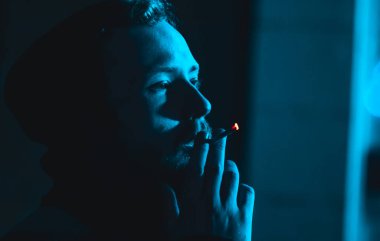 Karanlıkta kapüşonlu sigara içen yakışıklı bir adamın portresi soğuk mavi ışıkla 