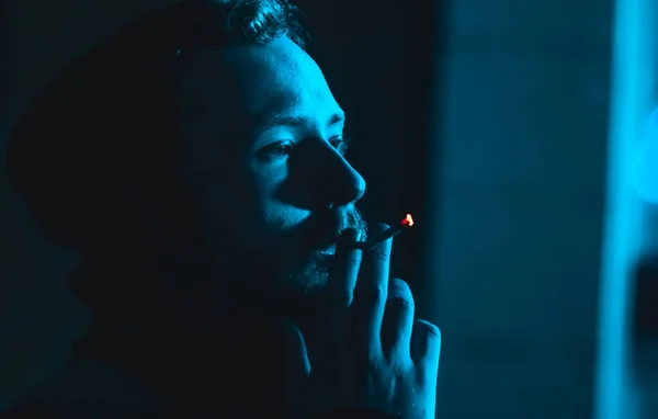 Portrait d'un bel homme fumeur dans une hotte à l'obscurité avec une lumière bleue froide Photo De Stock